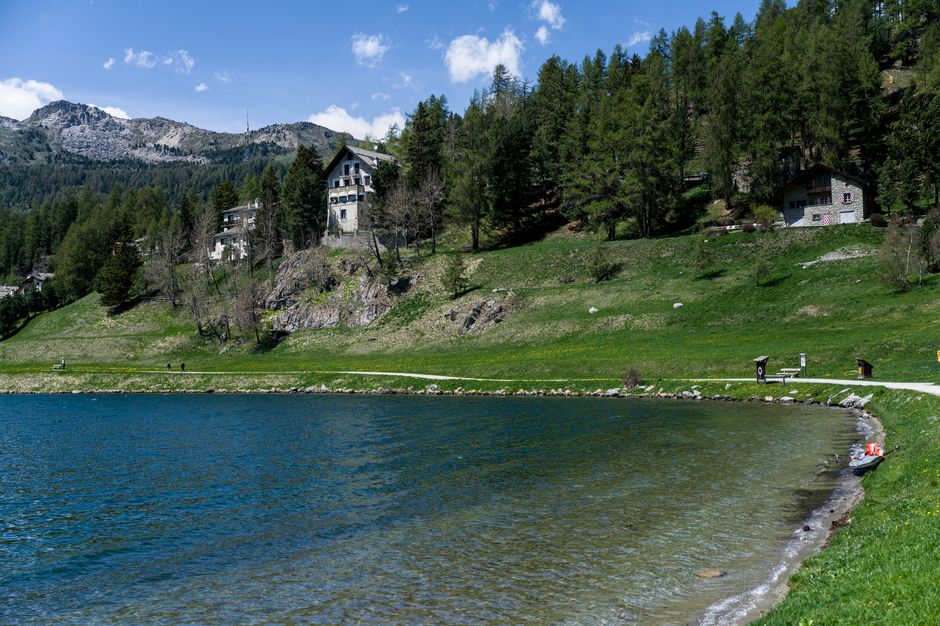 A scenic lakeside landscape featuring a calm, blue body of water with a gentle shoreline covered in grass and small rocks. On the edge of the water, there is a strip of sandy and pebbly beach, with a